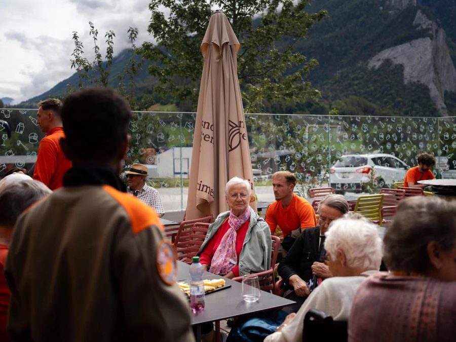 Résidents et bénévoles attablés en terrasse pour la pause de l'après-midi, montagnes en arrière-plan.